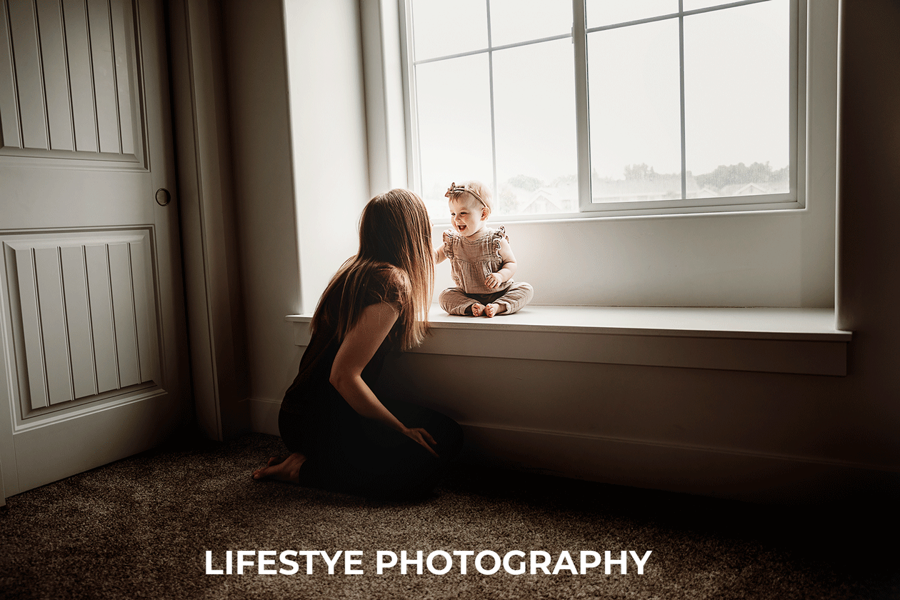 boise-best-family-photography-mom-with-long-hair-kneeling-near-window-bench-smiling-at-baby-girl-sitting-in-sunlight-smiling-big