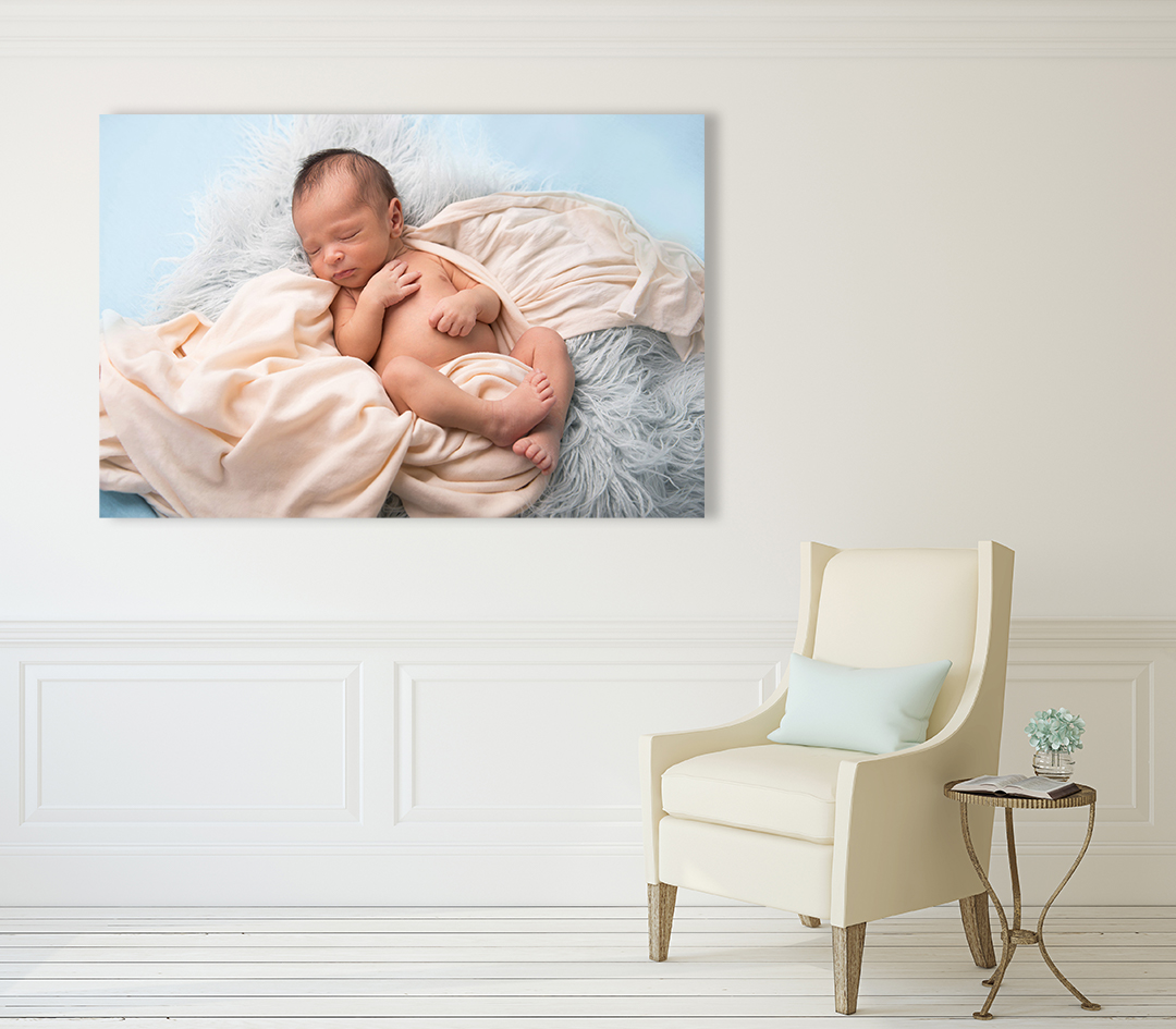 Beautiful canvas print of baby boy lying on cream blanket hanging on a cream wall with a brown chair and tiny table by the wall