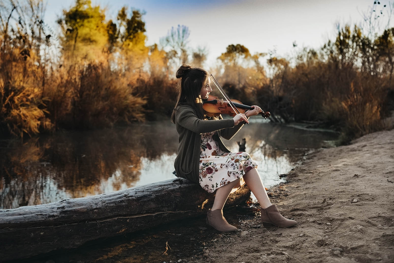 Young girl playing violin sitting on a log near water wearing a floral white dress with green cardigan