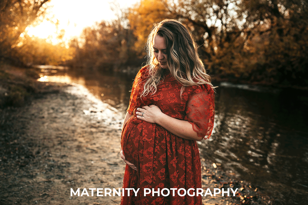 pregnant mama in red lace dress and blond hair looking down at her belly standing on the river