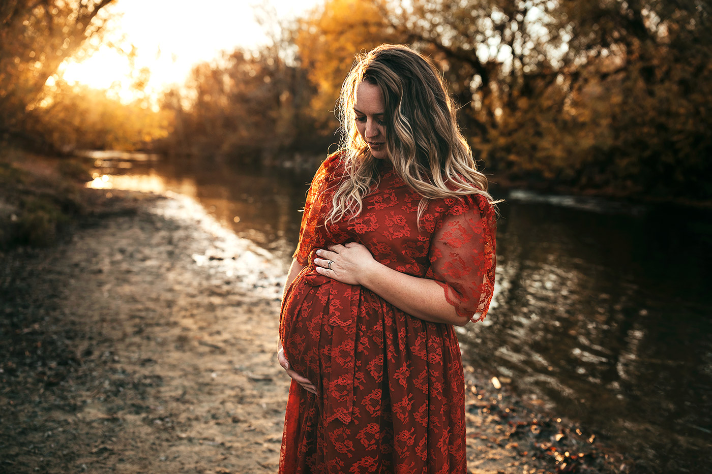 boise best maternity photographer beautiful blond pregnant mom in red lace dress holding her belly and looking at it standing on the river
