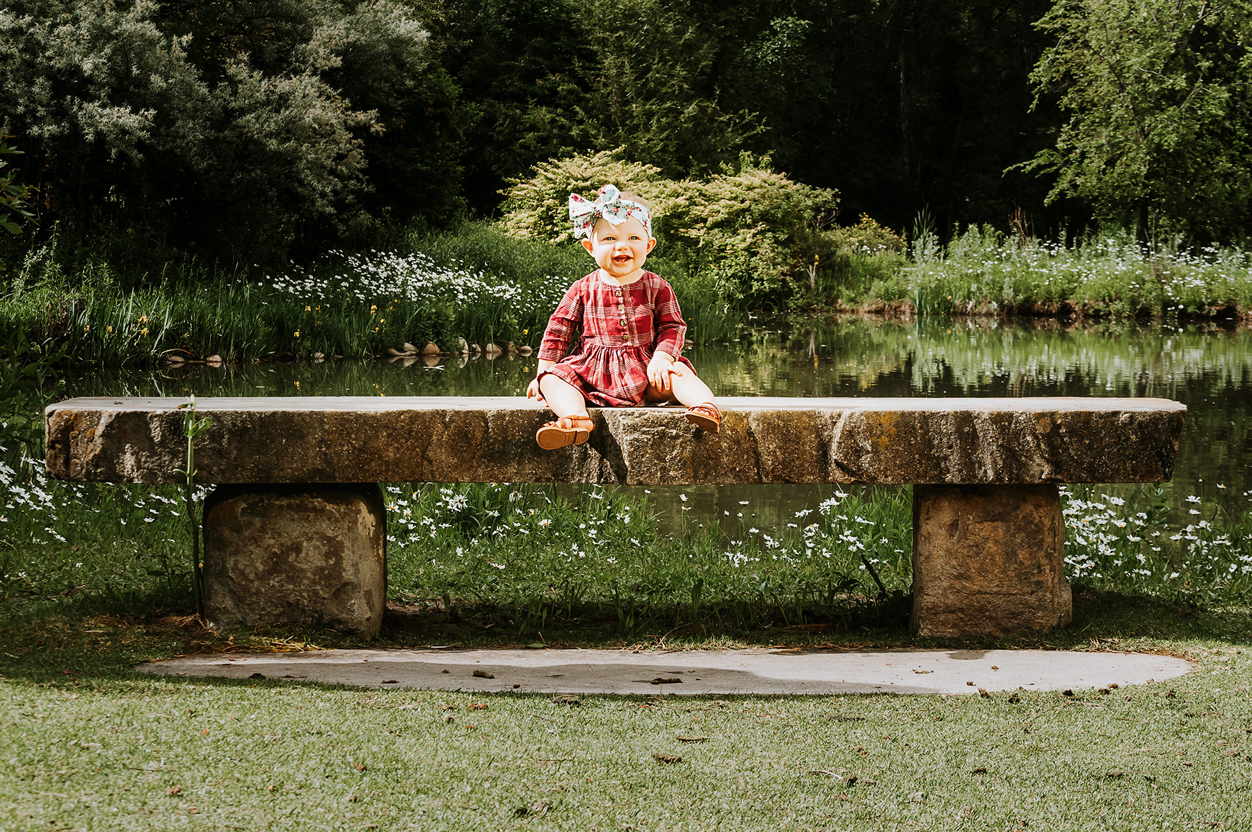 baby girl with maroon dress sitting on stone bench in front of pond