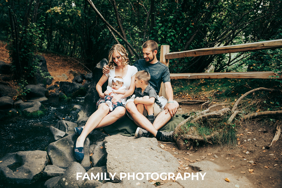 Family Photographer: family of four with baby sitting by creek looking at the baby