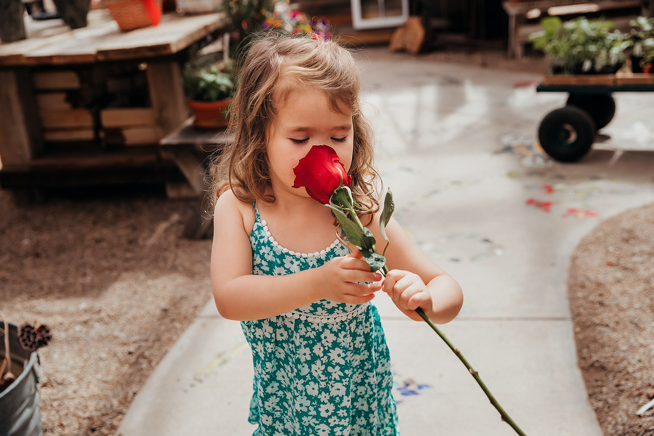 beautiful 4 year old girl in green dress in a greenhouse smelling a rose wearing
