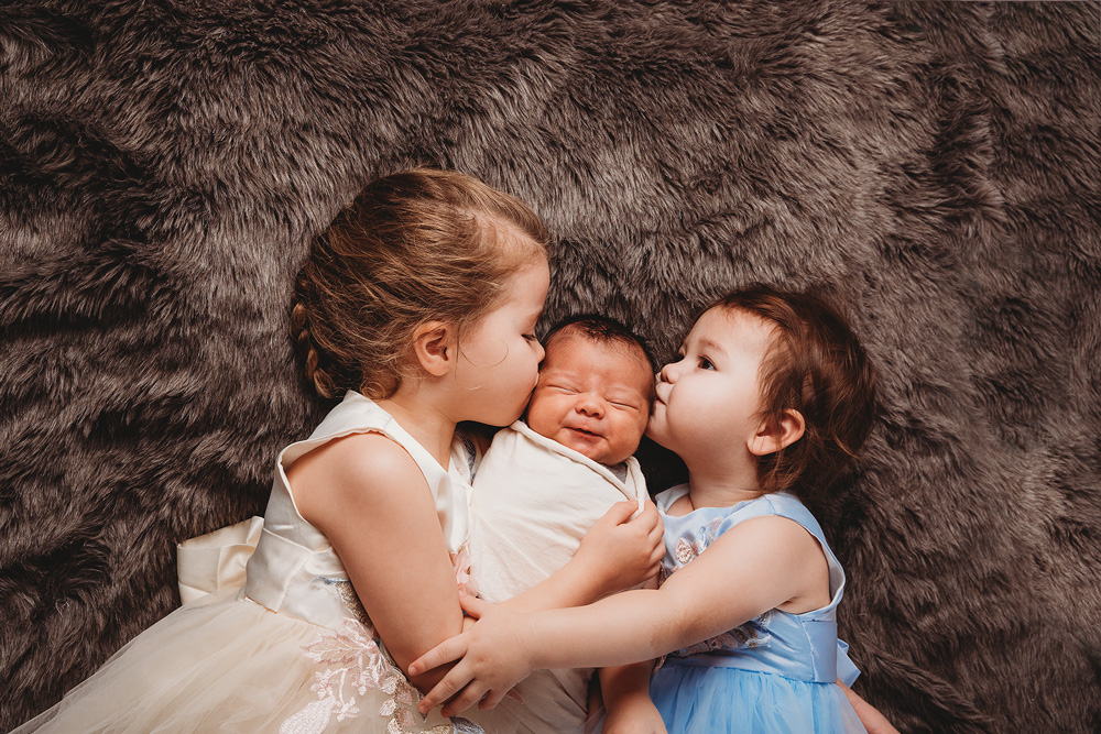 boise newborn photography two older sisters holding baby brother in the middle and giving him a big kiss